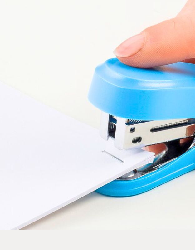 Close-up of a finger pressing down on a blue mini stapler stapling white paper